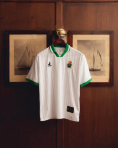 07-2web_400x_72715190-7259-4e3f-b989-821faf71e6ac | UAE SPORT STORE White polo shirt with green trim on collar and sleeves hanging on a wooden door, crest on the left chest and a small black brand logo on the right, framed sailboat pictures in the background