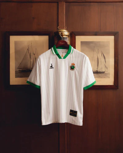 07-2web_400x_72715190-7259-4e3f-b989-821faf71e6ac | UAE SPORT STORE White polo shirt with green trim on collar and sleeves hanging on a wooden door, crest on the left chest and a small black brand logo on the right, framed sailboat pictures in the background