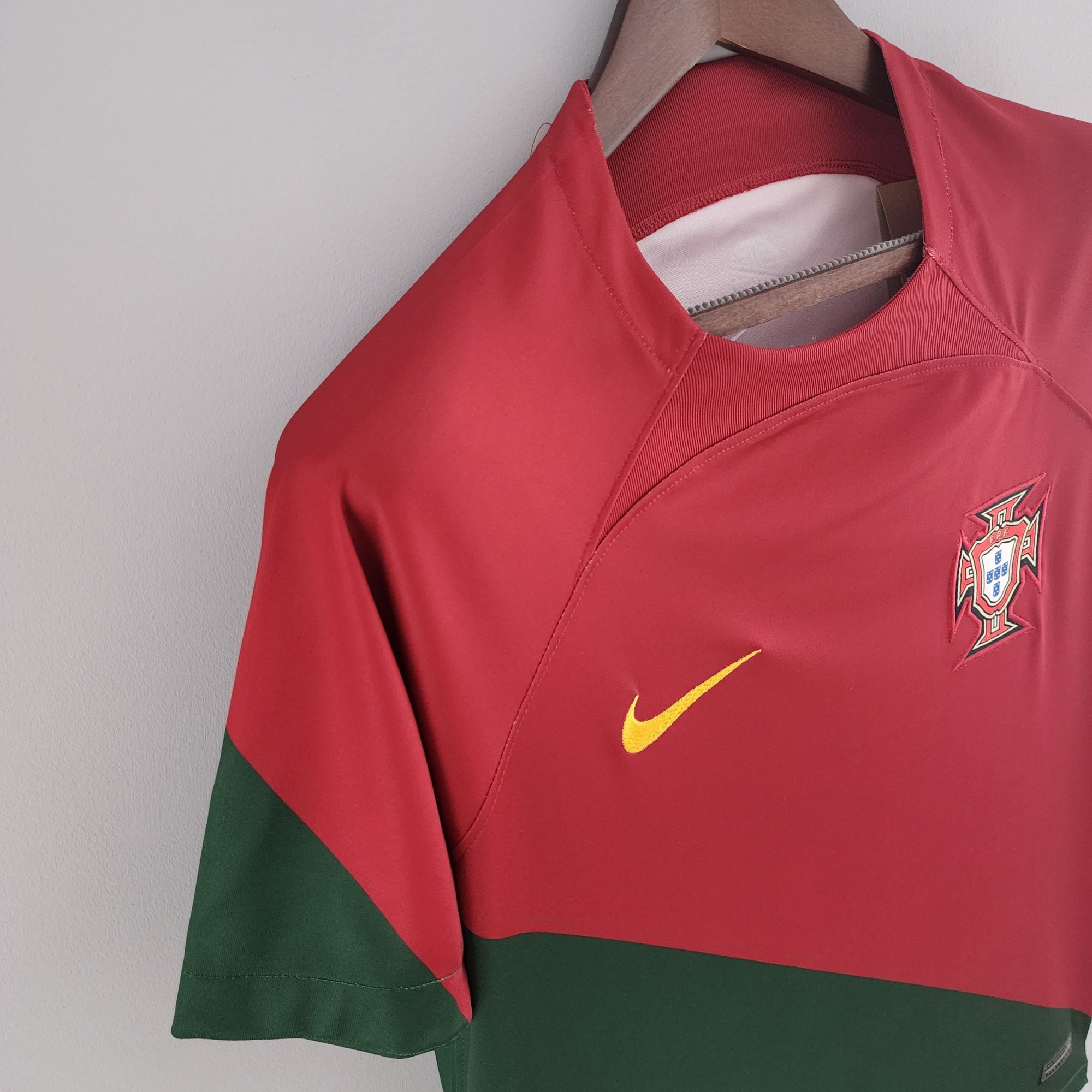 07175b34_92fa8f4d-3da1-47be-932a-6bbf7361a7ee | UAE SPORT STORE Red sports jacket hanging on a wooden hanger, with a crest badge on the chest and a yellow Nike swoosh nearby.