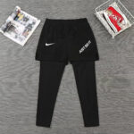 Black Nike athletic shorts layered over black leggings, featuring a white swoosh and 'JUST DO IT' text, laid flat on a gray wood floor with a magazine on the left and red sneakers in a white basket on the right.