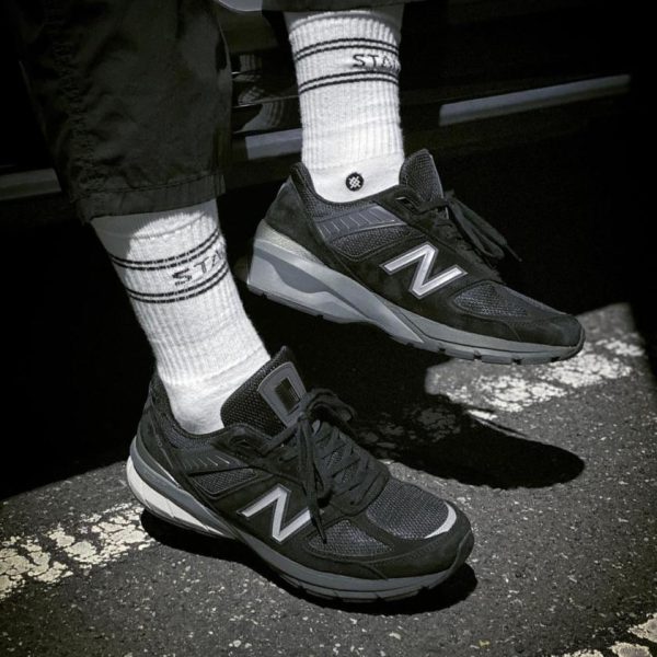 Close-up of black New Balance sneakers on white crew socks, standing on dark asphalt in front of a car.