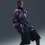 Male model wearing a navy England soccer jersey with red abstract pattern and crest, posing against a gray backdrop.