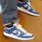 Blue sneakers with white laces and a purple Nike swoosh, worn with rolled jeans, on a wooden floor.