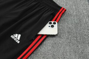 White iPhone with triple camera resting on black Adidas track pants with red stripes on a gray carpet, showing brand logo nearby.”