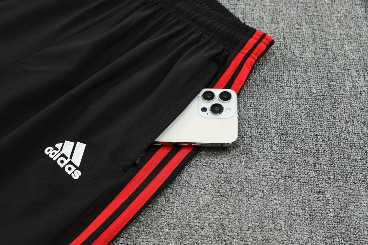 White iPhone with triple camera resting on black Adidas track pants with red stripes on a gray carpet, showing brand logo nearby.”