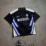 Inter Milan black away football jersey with blue and white diagonal stripes, Pirelli sponsor and Nike logo, laid on concrete with sneakers at the bottom