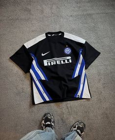 Inter Milan black away football jersey with blue and white diagonal stripes, Pirelli sponsor and Nike logo, laid on concrete with sneakers at the bottom