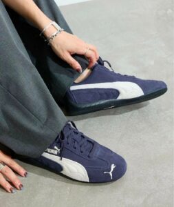 Close-up of a person wearing navy Puma sneakers with a white stripe, adjusting the laces while seated on a gray floor; hands visible with bracelets.