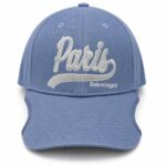 Blue baseball cap with white embroidered 'Paris' script and 'Balenciaga' logo on the front panel; curved brim and structured crown.