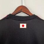Black garment on a wooden hanger, close-up of the collar against a light wall; small badge with a red circle is centered on the chest area.