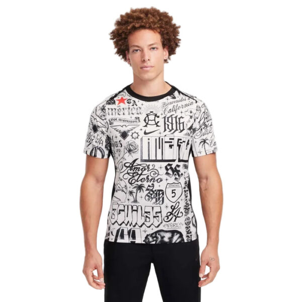 Young man modeling a white graphic T-shirt with black tattoo-style text and designs, paired with black pants