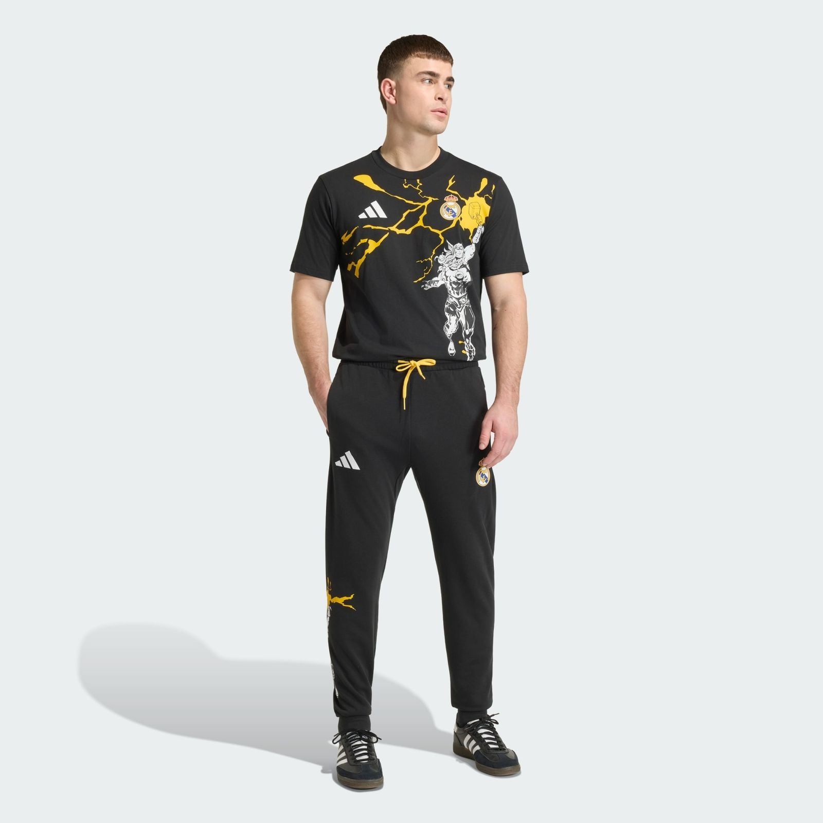 09dc22a4-c585-4883-a10b-0e163a08714d | UAE SPORT STORE Man standing in a black Real Madrid training outfit with yellow lightning graphics and branding logos (Adidas and Real Madrid crest).