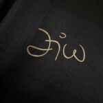 Gold cursive signature on dark textured fabric as a close-up detail.