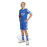 Young boy in Real Madrid blue football kit, hands on hips, posing in white sneakers.