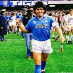 Soccer player in a blue Mars jersey walks on the field, fist raised, with fans and a cameraman in the background.