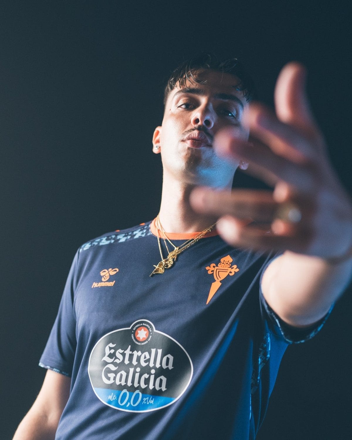 Portrait of a young man in a navy blue sports jersey with Estrella Galicia logo, reaching toward the camera with a focused expression.