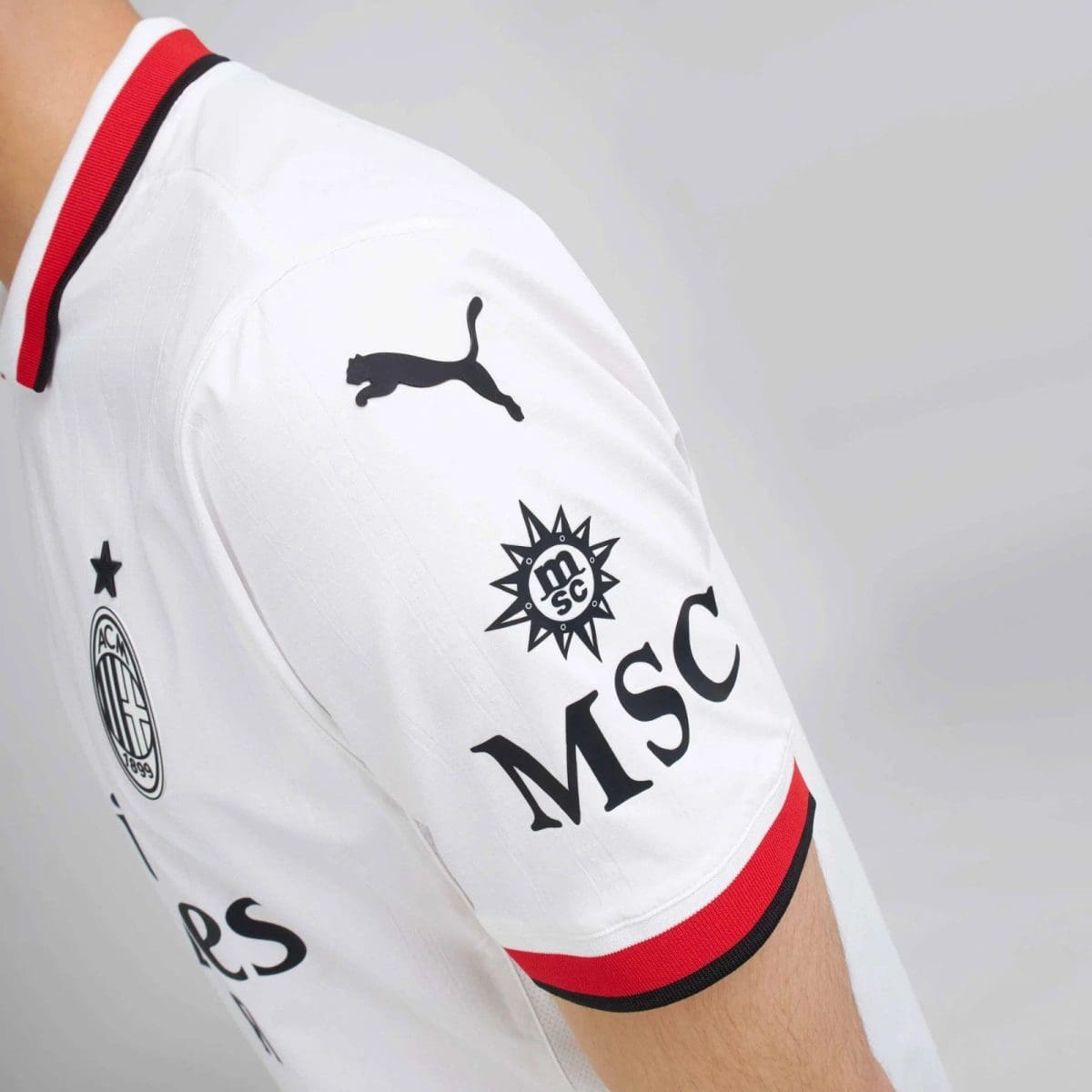 White soccer jersey sleeve with red and black trim, Puma logo and club crest visible on the arm sleeve, sponsor text beneath.