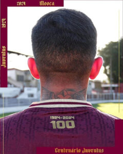 Back view of a person in a maroon jersey with '1924-2024' and '100' on the back; visible neck tattoo, outdoor field, commemorative frame text nearby.
