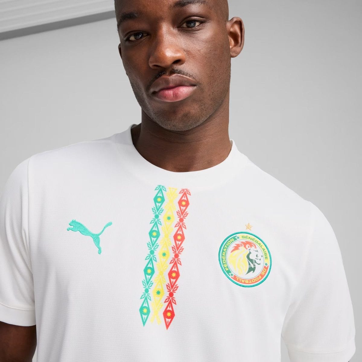 Close-up of a man wearing a white jersey featuring the Senegal Football Federation crest and multicolored vertical patterns in the center of the chest area.