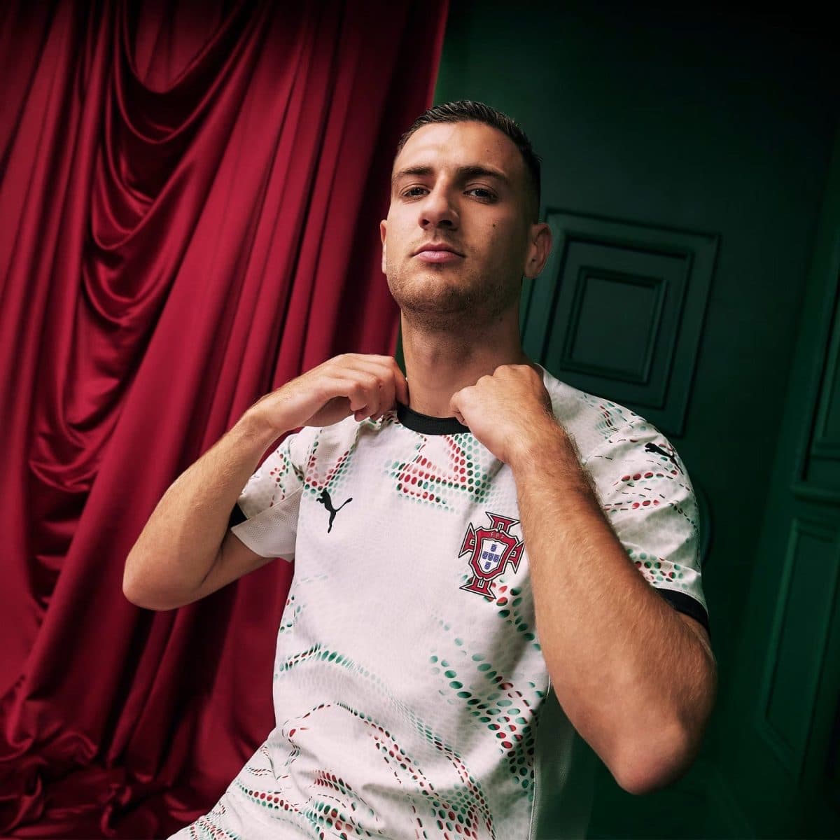 Man in Portugal national team jersey adjusts his collar, posed against a red curtain on the left and a green wall on the right.