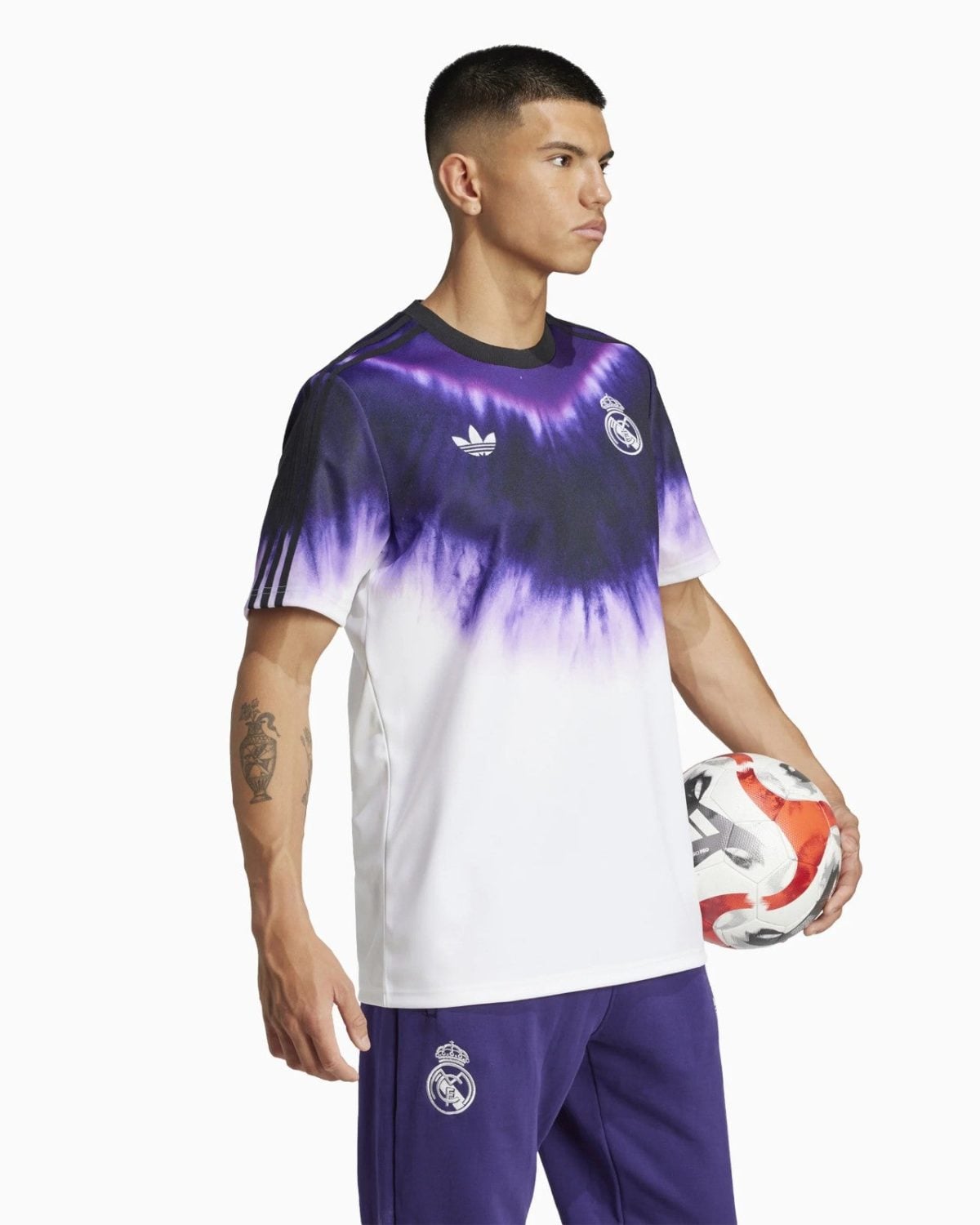 Male soccer player in a tie-dye purple and white Real Madrid jersey, holding a soccer ball at his side, against a white background.
