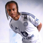 Footballer in a white Leicester City kit, leaning forward toward the camera with a focused expression.