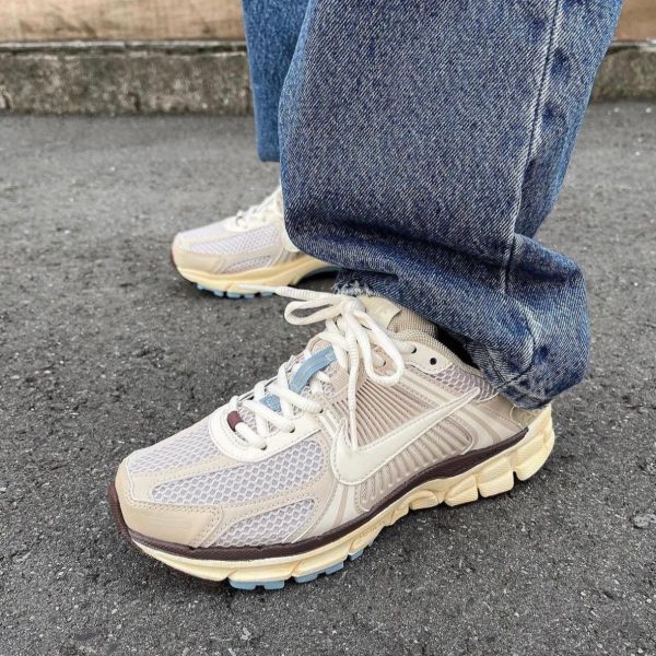 Cream-colored running shoes with white laces and mesh panels, worn with cuffed blue jeans on a paved surface.