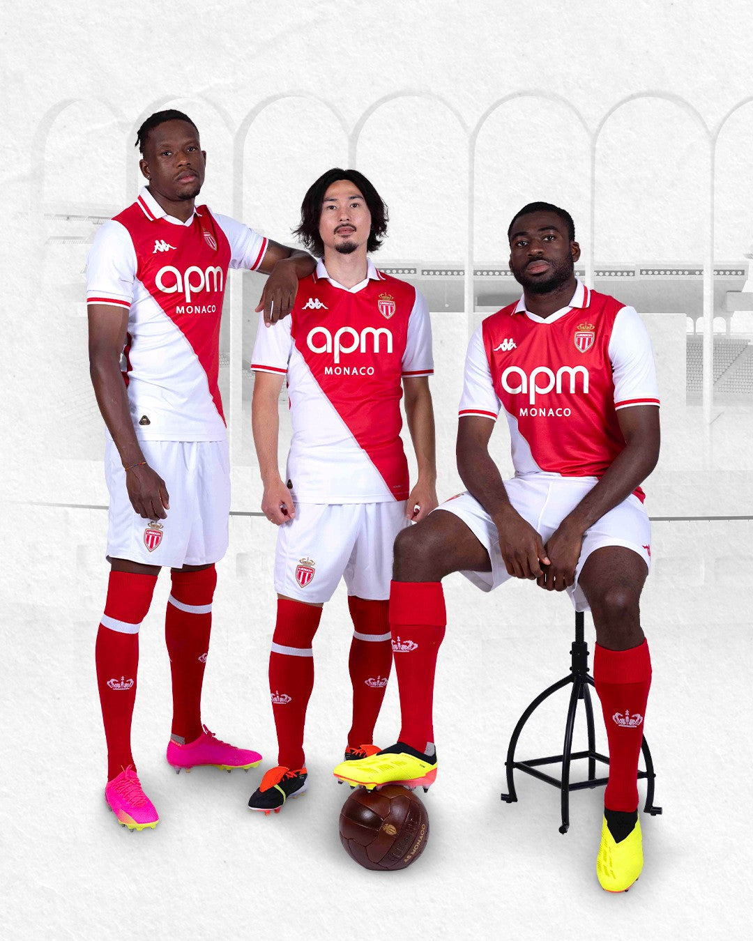 0a5yIbLod5PgzOQ | UAE SPORT STORE Three AS Monaco soccer players in red-and-white uniforms pose together, one seated on a stool with a soccer ball at their feet and two standing beside him in a stadium backdrop.