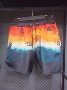 Colorful men's swim trunks with an orange-to-blue gradient and repeating logos, hanging on a hanger on a display rack.