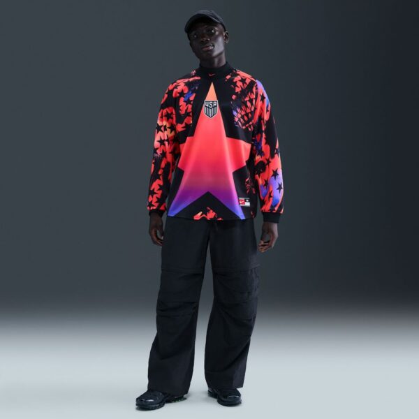 Person wearing a bright star-patterned USA soccer training jacket with a black cap and black cargo pants, standing against a dark backdrop.