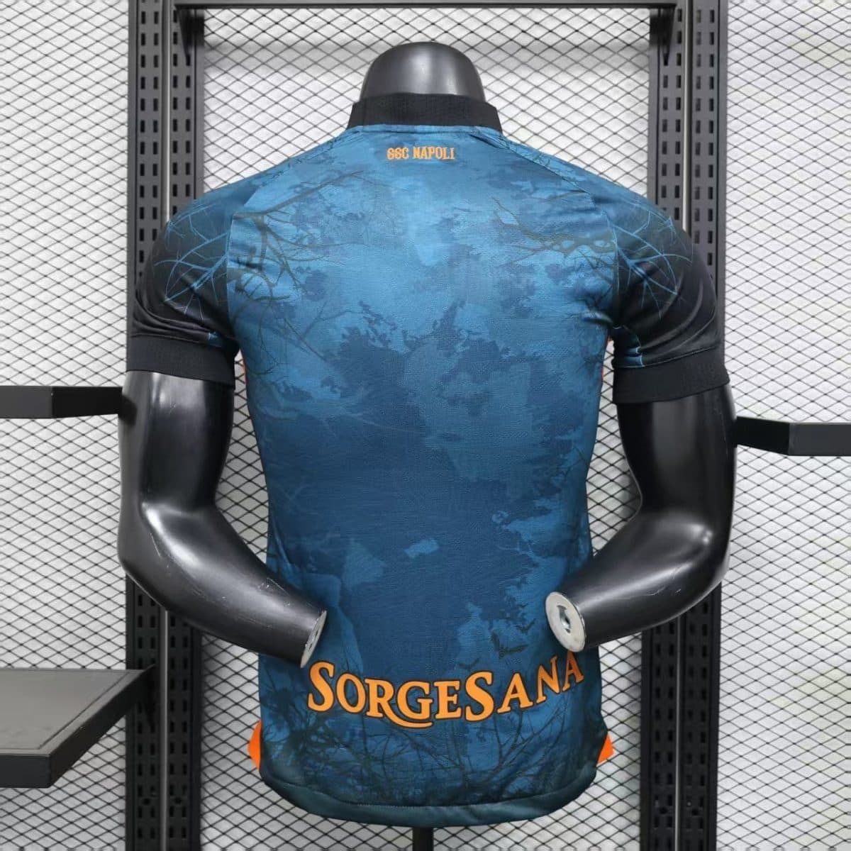 Back view of a blue patterned sports jersey on a mannequin, with orange text near the lower back reading SORGESANA and small orange text SSC NAPOLI at the neck area.