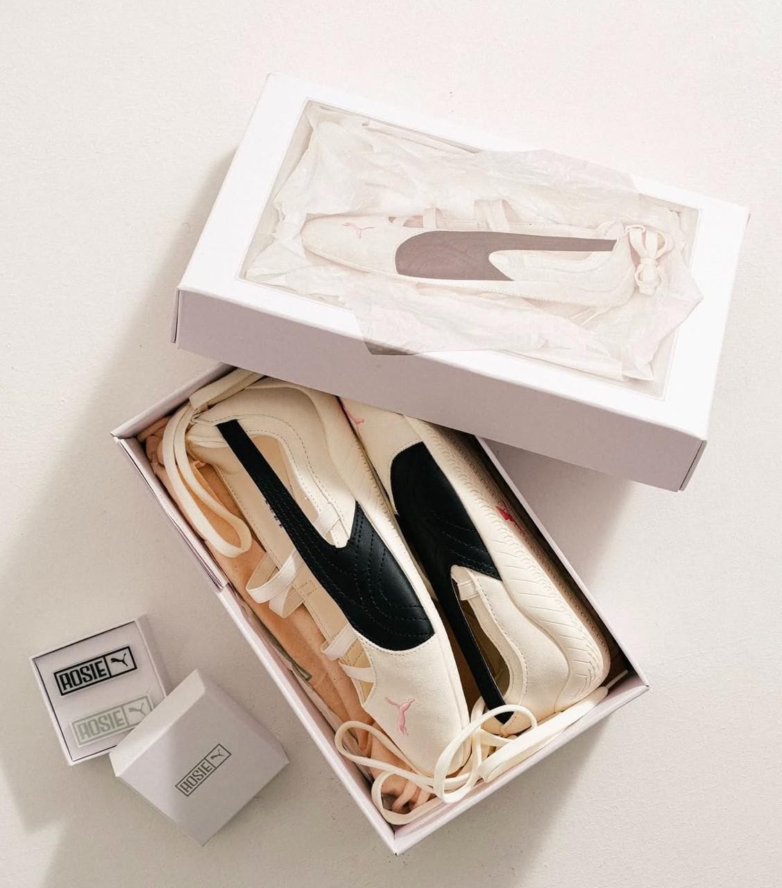 0b2bc439-fdad-4d7b-8004-cecc423f2d93 | UAE SPORT STORE Open white shoebox with tissue paper reveals cream-colored sneakers with a black side panel and pink stitching details; brand Rosie visible on inserts nearby.