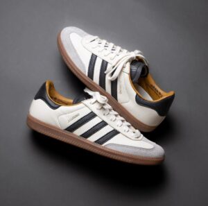 White Adidas-style sneakers with black stripes and a gum brown sole on a gray background.