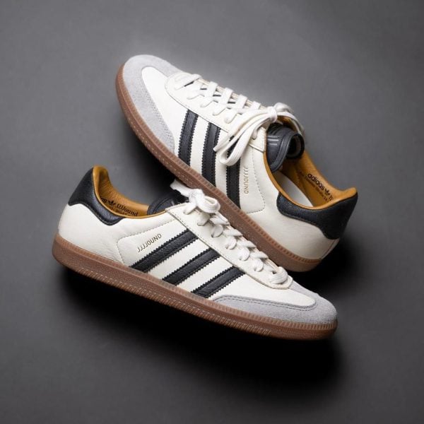 White Adidas-style sneakers with black stripes and a gum brown sole on a gray background.