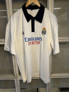 White Real Madrid polo jersey on a hanger with black collar, featuring Emirates Fly Better sponsor and club crest on the chest, Adidas logo on the sleeve, hanging in a display case.
