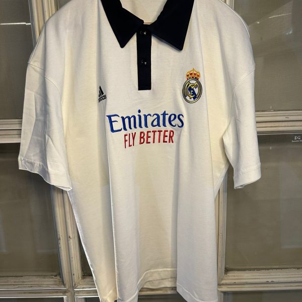 White Real Madrid polo jersey on a hanger with black collar, featuring Emirates Fly Better sponsor and club crest on the chest, Adidas logo on the sleeve, hanging in a display case.