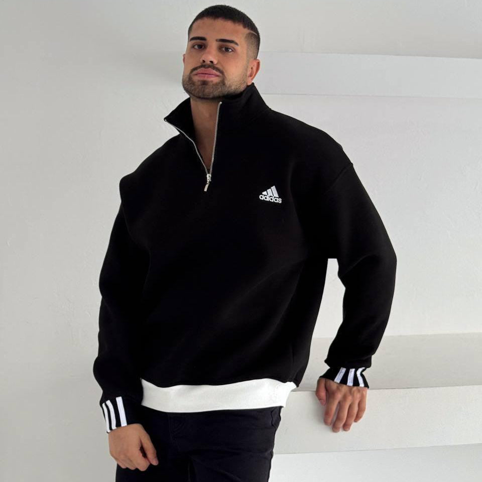 0beb98ad-e177-4e03-ac08-c86705c053ea Man wearing a black Adidas quarter-zip fleece with a white hem, posing against a light wall.