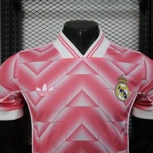 Front view of a pink Real Madrid football jersey with the club crest and Adidas logo on a geometric pattern.