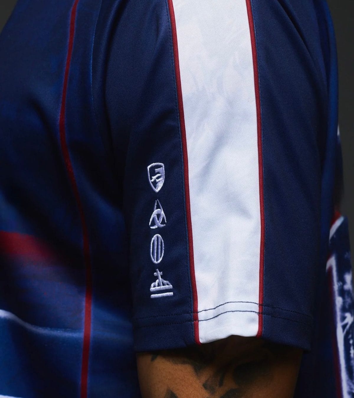Close-up of a navy blue sports jersey sleeve with a white center stripe bordered in red and small white embroidered symbols along the seam.