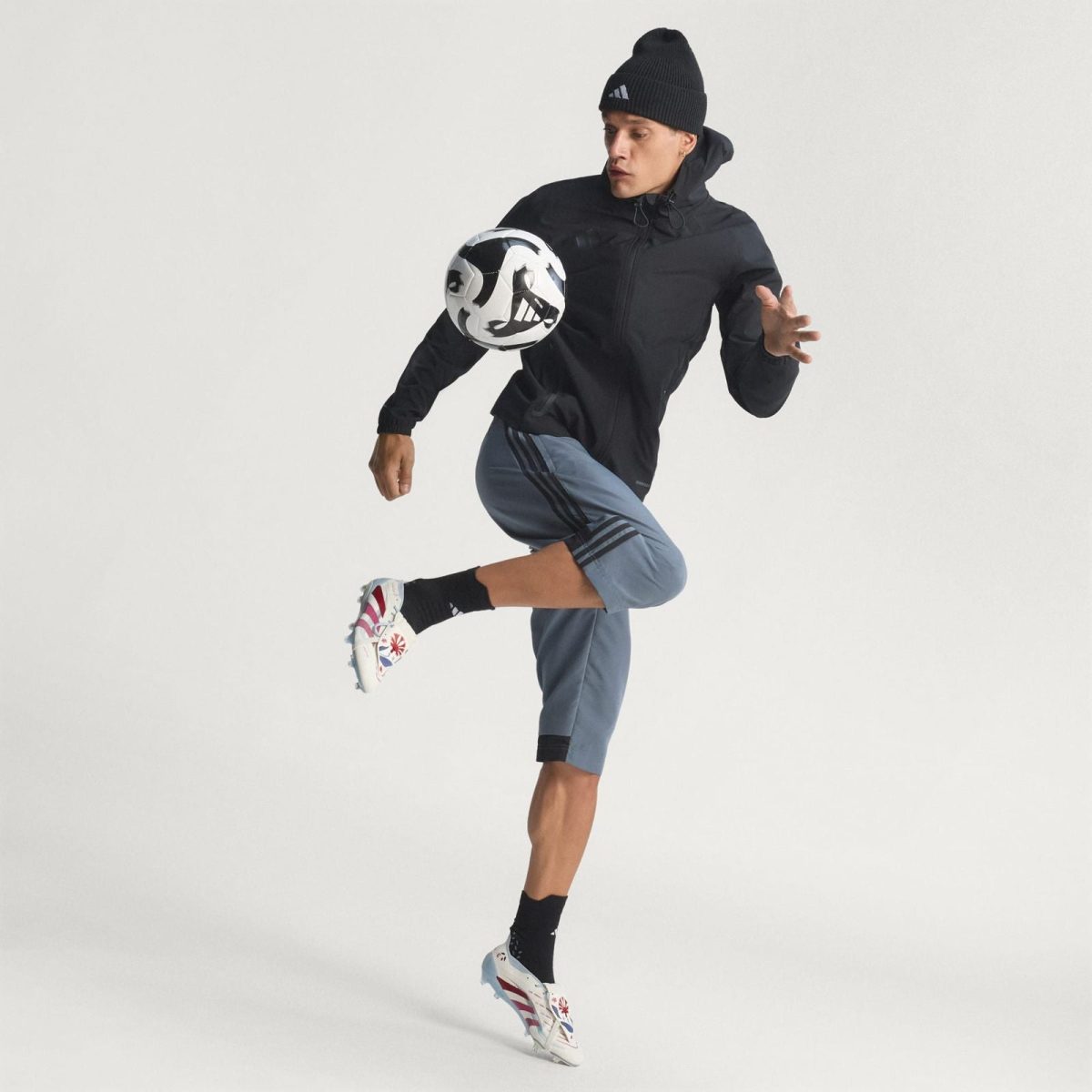 Athlete in a black jacket and beanie kicking a soccer ball midair
