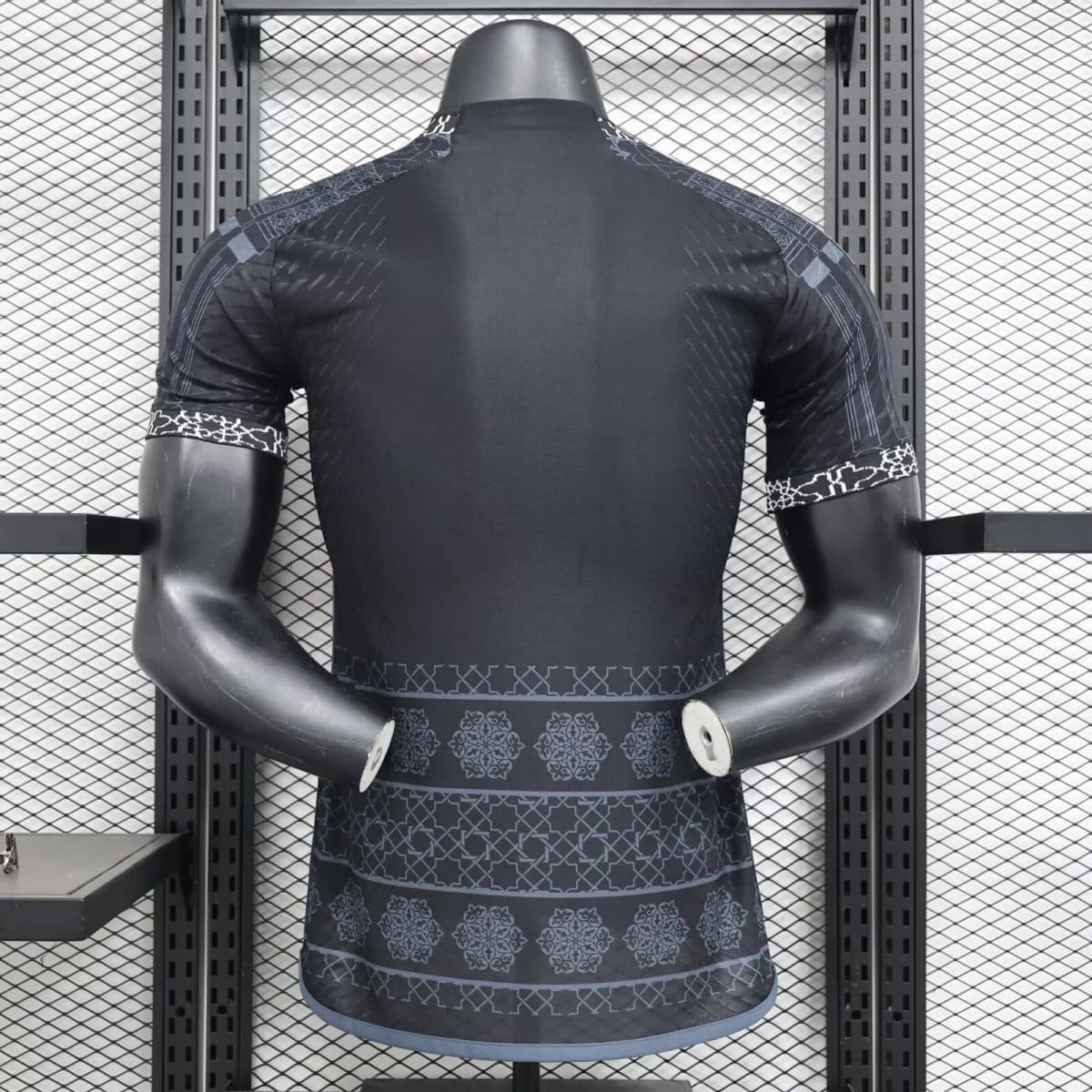 Back view of a dark athletic shirt on a mannequin in a metal rack, showcasing patterned sleeves and hem in a gym display.