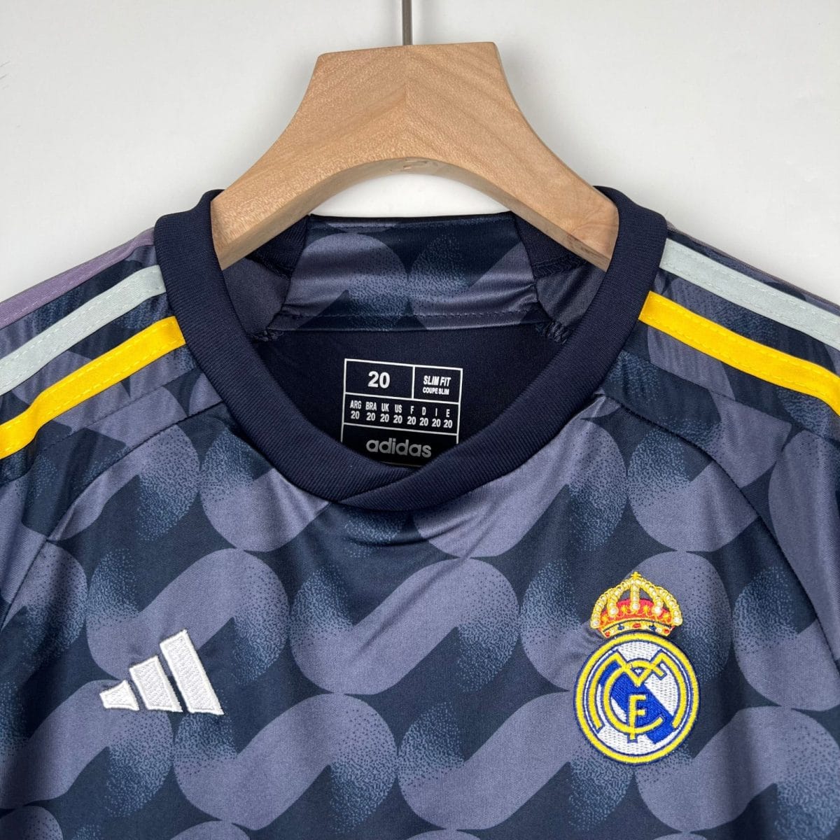 Close-up of a Real Madrid football jersey on a wooden hanger, showing the Adidas logo and Real Madrid crest with navy and camouflage pattern and yellow accents.