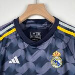 Close-up of a Real Madrid football jersey on a wooden hanger, showing the Adidas logo and Real Madrid crest with navy and camouflage pattern and yellow accents.