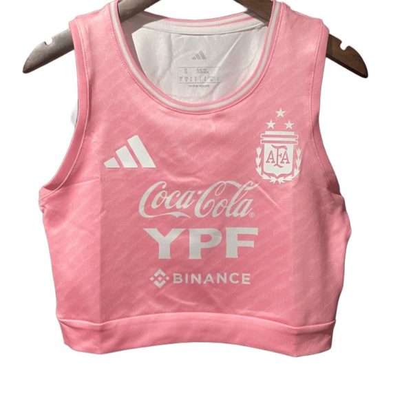 Pink cropped athletic top with Adidas logo, Argentina crest, and sponsor logos (Coca-Cola, YPF, Binance) on chest, hanging on a wooden hanger in front of a white background.