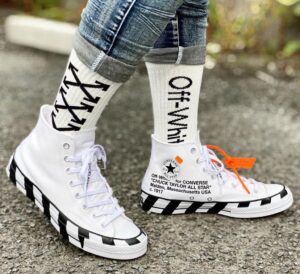 White high-top sneakers with a black diagonal stripe sole on pavement, worn with rolled denim and bold Off-White socks with an orange zip-tie tag.