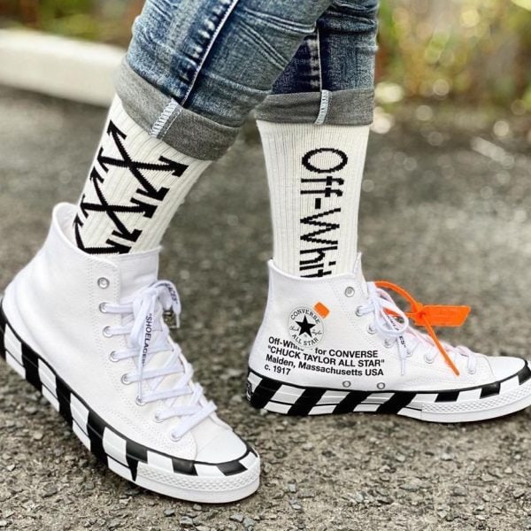 White high-top sneakers with a black diagonal stripe sole on pavement, worn with rolled denim and bold Off-White socks with an orange zip-tie tag.