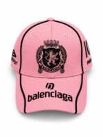 Pink Balenciaga baseball cap with a black crest emblem on the front and black outlined stitching along the brim, plus side logos.