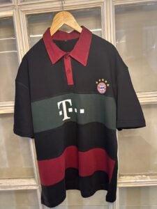 Oversized black polo with red collar and horizontal color blocks, Bayern Munich crest on the chest, hanging on a wooden hanger.