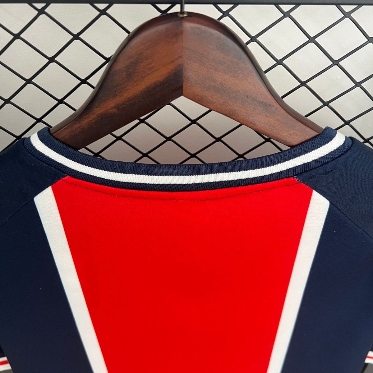 Red athletic jersey with white and navy trim hung on a wooden hanger in front of a chain-link fence in the background.