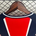 Red athletic jersey with white and navy trim hung on a wooden hanger in front of a chain-link fence in the background.